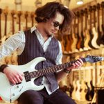 Handsome stylish man in classical vest and sunglasses smiling while playing an electric guitar in a musical shop